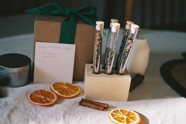 Tee als Geschenk: Glasröhrchen mit losem Tee in einem Holzständer neben einer Geschenkbox mit grünem Band