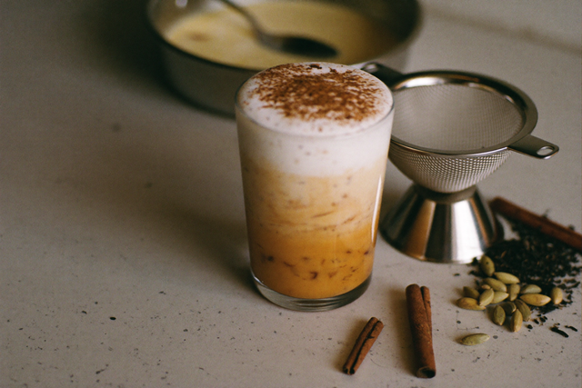 Glas Chai Latte mit Milchschaum und Zimt neben einem Teesieb mit losem Chai-Tee